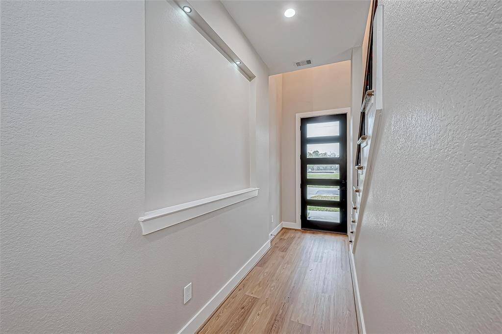 This photo shows a bright and modern hallway with light wood flooring and recessed lighting. The space leads to a sleek, glass-paneled front door, creating a welcoming entryway.