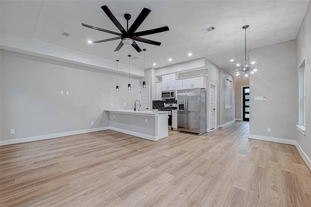 This photo showcases a modern open-concept living area with sleek hardwood floors, a spacious kitchen featuring stainless steel appliances, white cabinetry, and an island with pendant lighting. A large ceiling fan and unique light fixtures add style, while ample natural light enhances the airy feel.