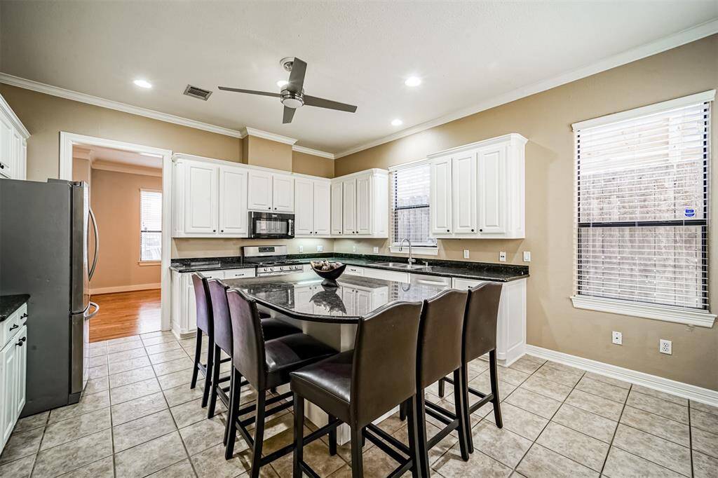 This kitchen features a large island with seating, granite countertops, and stainless steel appliances. It has ample cabinet space, a ceiling fan, and plenty of natural light.