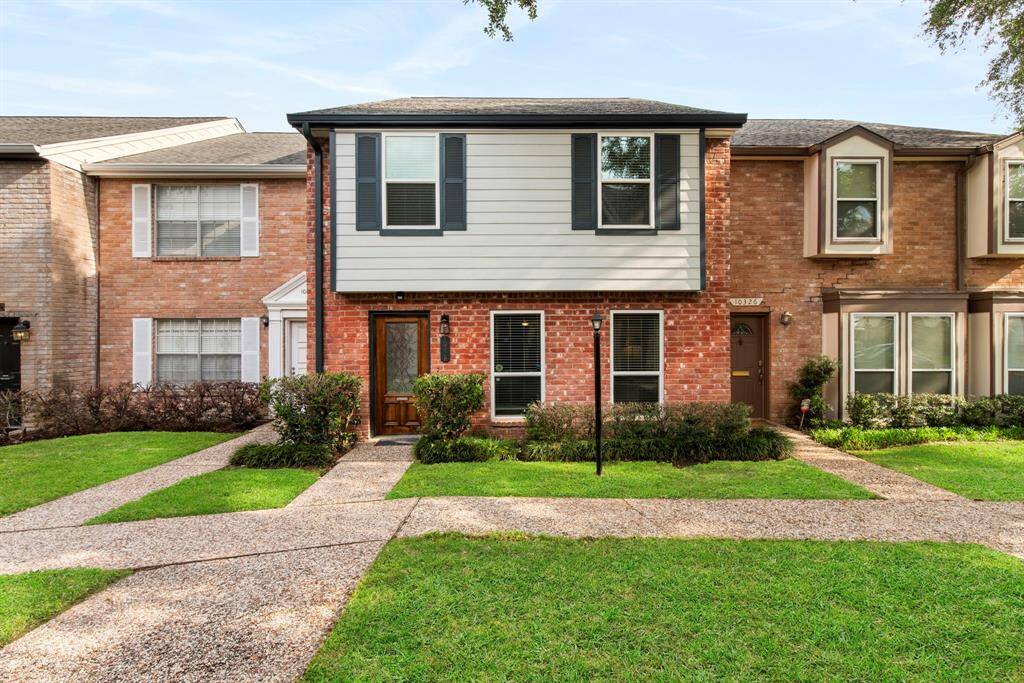 Charming red brick exterior with black shutters and a carved wood front door, framed by neat hedges and a welcoming walkway.