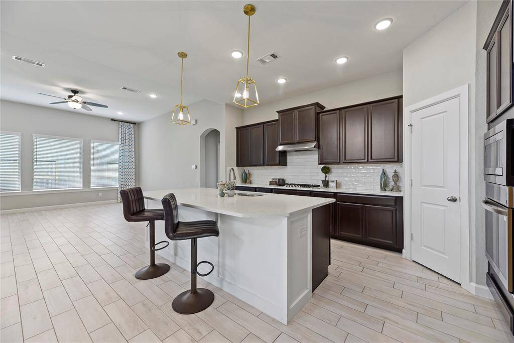 This modern kitchen features dark wood cabinets, a sleek white island with seating, and stainless steel appliances and upgraded pendant lighting. It opens to a spacious living area with large windows and light wood flooring, creating a bright, inviting space.