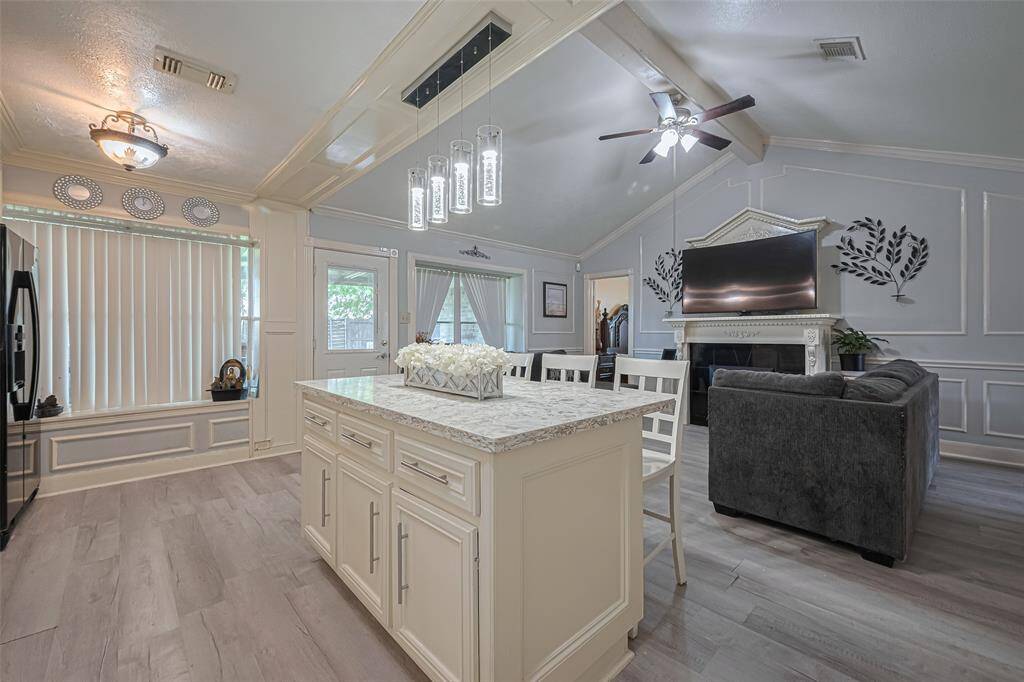 This spacious and elegant living area features a bright, open layout with a modern kitchen island, high ceilings, and a cozy faux fireplace. The large windows allow natural light to fill the room, creating a warm and inviting ambiance.