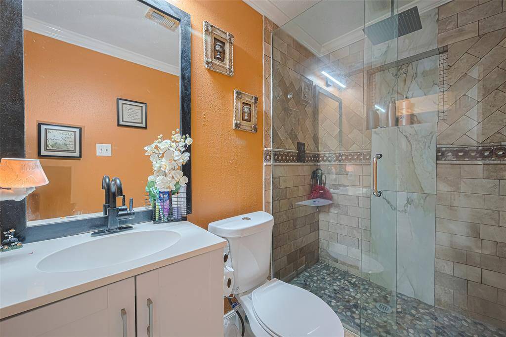 This bathroom features a stylish walk-in shower with natural stone flooring and elegant tile work. It has a modern vanity with a large mirror, vibrant orange walls, and decorative accents, creating a warm and inviting space.