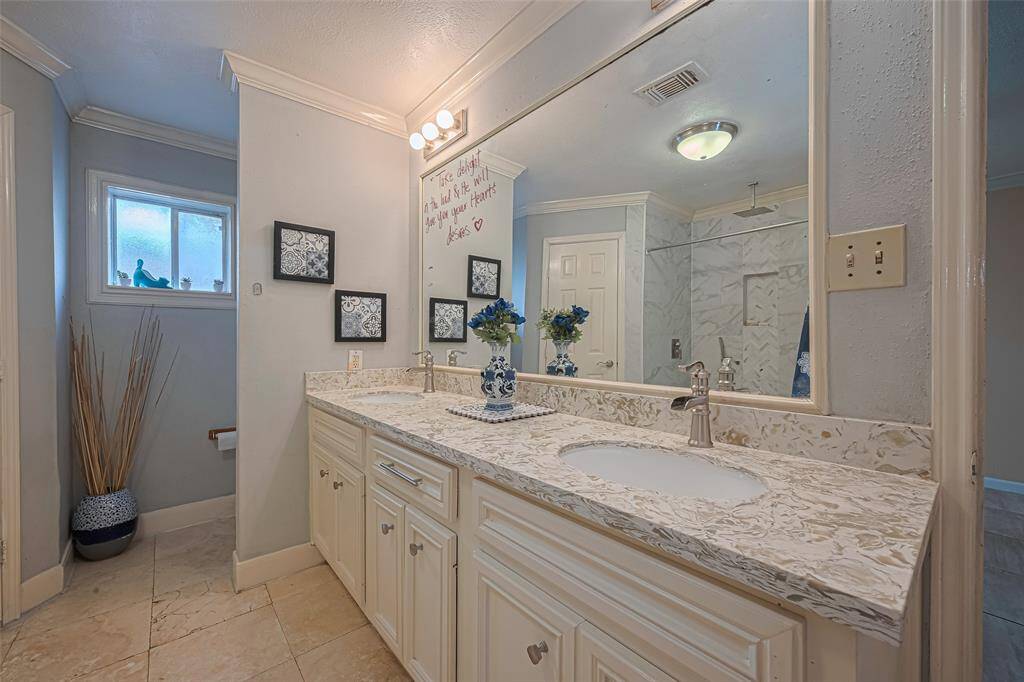 A dual-sink vanity with a marble countertop, large mirror, and elegant lighting. The space includes a frosted window for natural light, decorative touches, and a shower area with modern fixtures. Neutral tones add a calming atmosphere.