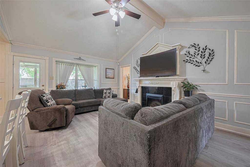 Cozy living room with vaulted ceilings, featuring a large sectional sofa and recliner. The space is brightened by natural light through a window and door, and centered around a decorative fireplace with a mounted TV. Elegant wall moldings and modern gray tones enhance the room's charm.