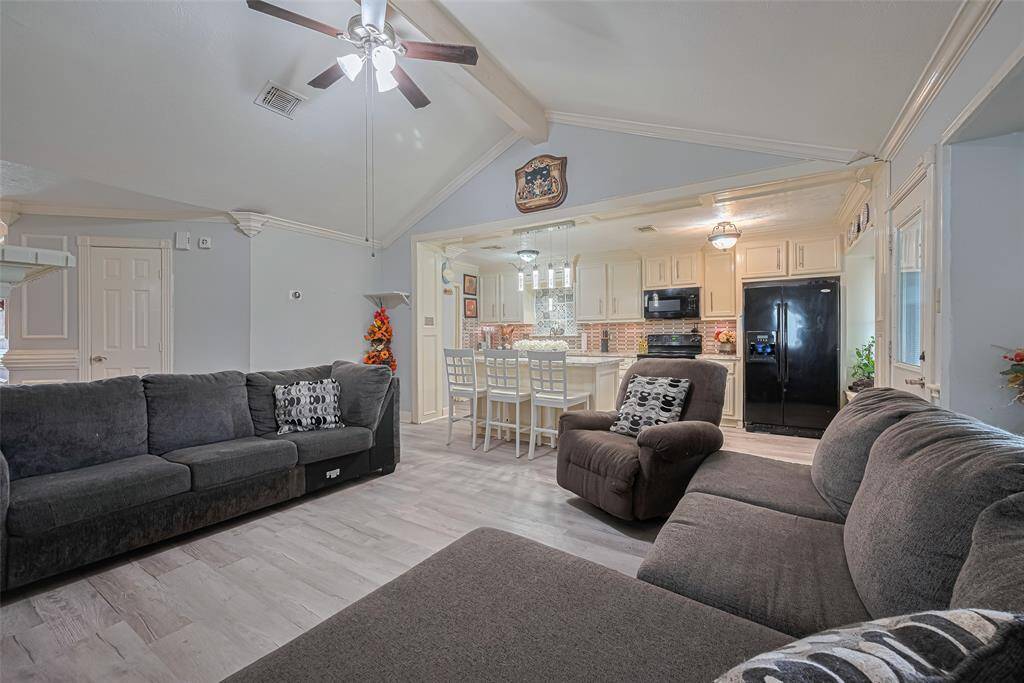 The room features a cozy seating arrangement, light wood flooring, and a ceiling fan. The adjacent kitchen is equipped with white cabinetry, a large island with bar seating, and black appliances, creating a stylish and functional space perfect for entertaining.