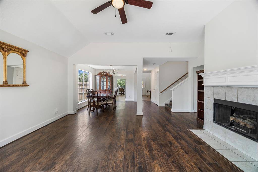 This photo showcases a spacious, open-plan family and dining area with hardwood floors, a ceiling fan, and a cozy fireplace. Natural light fills the room from large windows, and there's a view of a staircase leading to the upper level.
