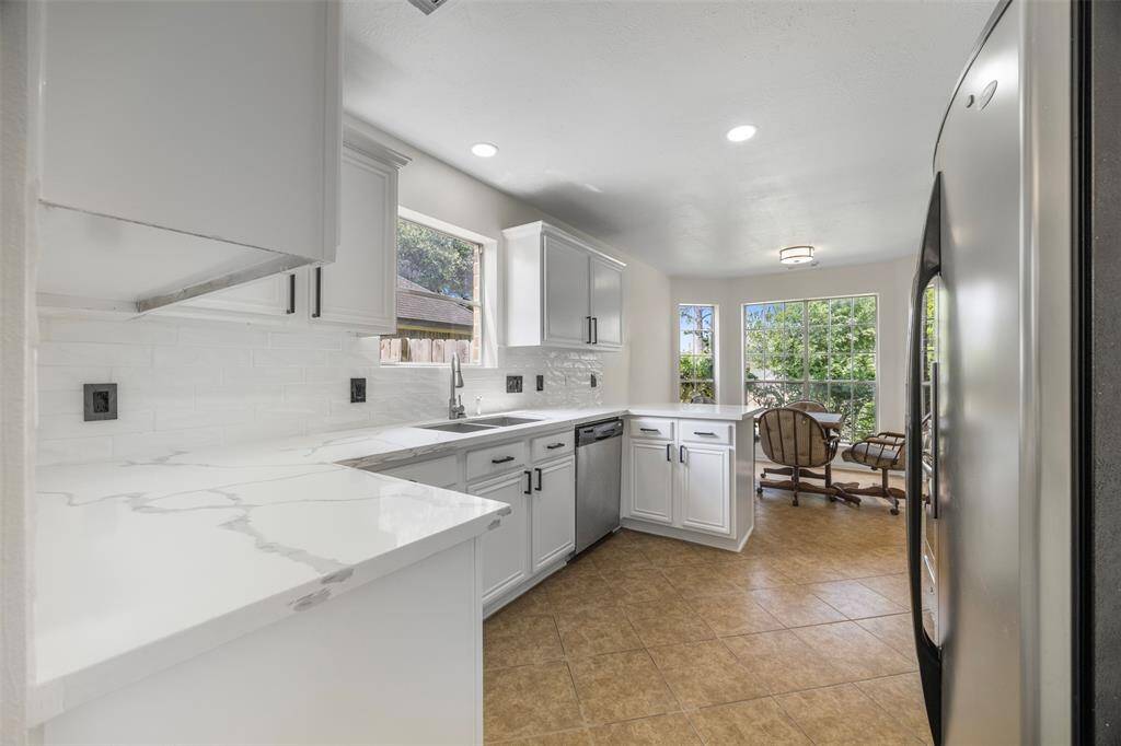 This photo showcases a bright, modern kitchen with white cabinetry and marble countertops. It features a stainless steel refrigerator, dishwasher, and a cozy dining area with large windows providing ample natural light. The space has a clean, open feel with tiled flooring.
