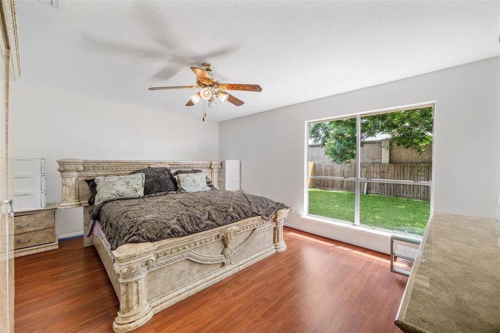 This primary bedroom features warm wood flooring, a ceiling fan and a large window offering plenty of natural light and a view of the fenced backyard, creating a cozy and inviting atmosphere.