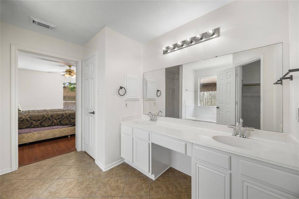 This photo showcases a bright bathroom with a double vanity, large mirror, and modern lighting. It features a tile floor and connects to primary bedroom with wood flooring and a ceiling fan. There's also a glimpse of a shower area and a window providing natural light.