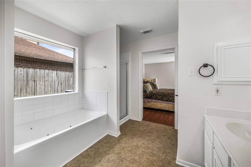 This bathroom features a spacious tub with jets, a large window for natural light, a separate shower, and direct access to a bedroom. Neutral tile flooring and white cabinetry create a clean, modern look.
