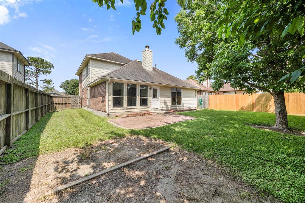 This photo shows a spacious backyard with a mix of grass and a patio area, surrounded by a wooden fence for privacy. The rear of a two-story house features large windows and a chimney, with mature trees providing shade.