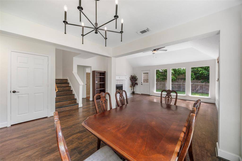 Bright and spacious dining area with hardwood floors, a modern chandelier, and large windows providing ample natural light. Open layout leads to a cozy living space with a fireplace and views of a fenced backyard. Adjacent staircase adds character.