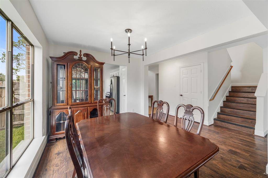 This photo showcases dining area featuring a large window, and modern lighting. The space connects to a kitchen through an open doorway and a staircase leading upstairs, all set on rich hardwood floors.