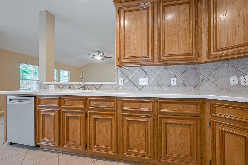 This kitchen features warm wooden cabinetry, a sleek countertop, and a modern dishwasher. The open layout connects to a living area with large windows and a ceiling fan, providing a bright and airy feel.