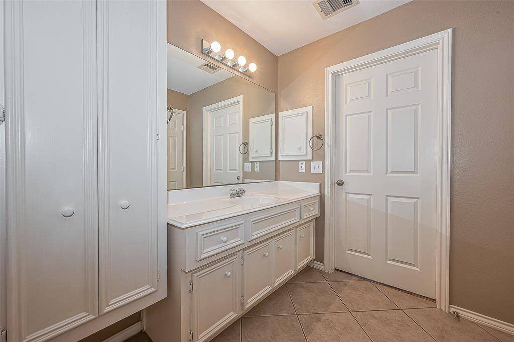 This bathroom features a clean, neutral design with light-colored walls and cabinetry. It includes a large mirror, ample countertop space, and a combination of under-sink and wall storage. The tiled floor and well-lit vanity area add to its functionality and appeal.