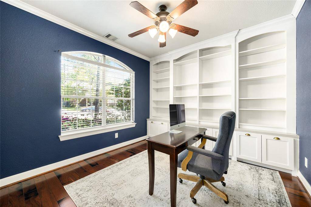 Bright home office with hardwood floors, featuring built-in white bookshelves and a large arched window. Ideal for a productive work-from-home setup.