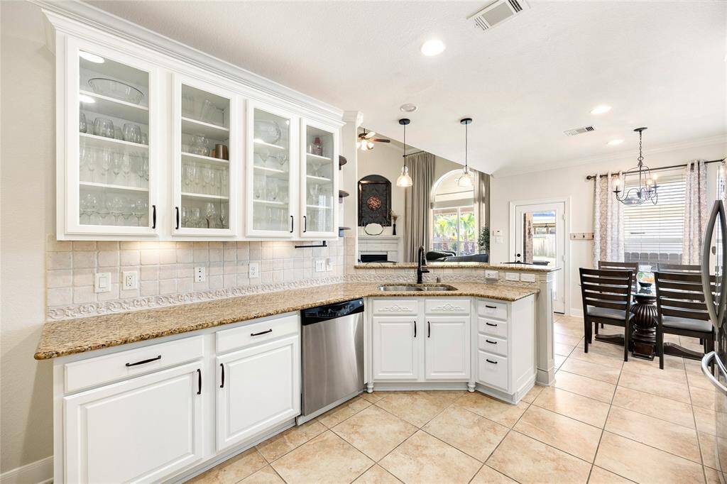 This photo showcases a bright, open-plan kitchen and dining area. The kitchen features white cabinetry with glass doors. Pendant lighting hangs over a central island with a sink. The dining area is adjacent, with large windows and a chandelier, providing a cozy yet spacious feel.