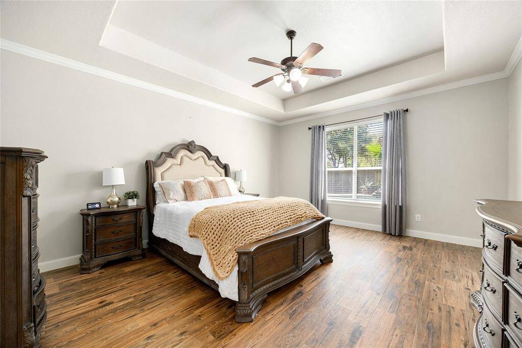 This spacious bedroom features elegant wood flooring, a stylish tray ceiling with a ceiling fan, and a large window offering plenty of natural light.