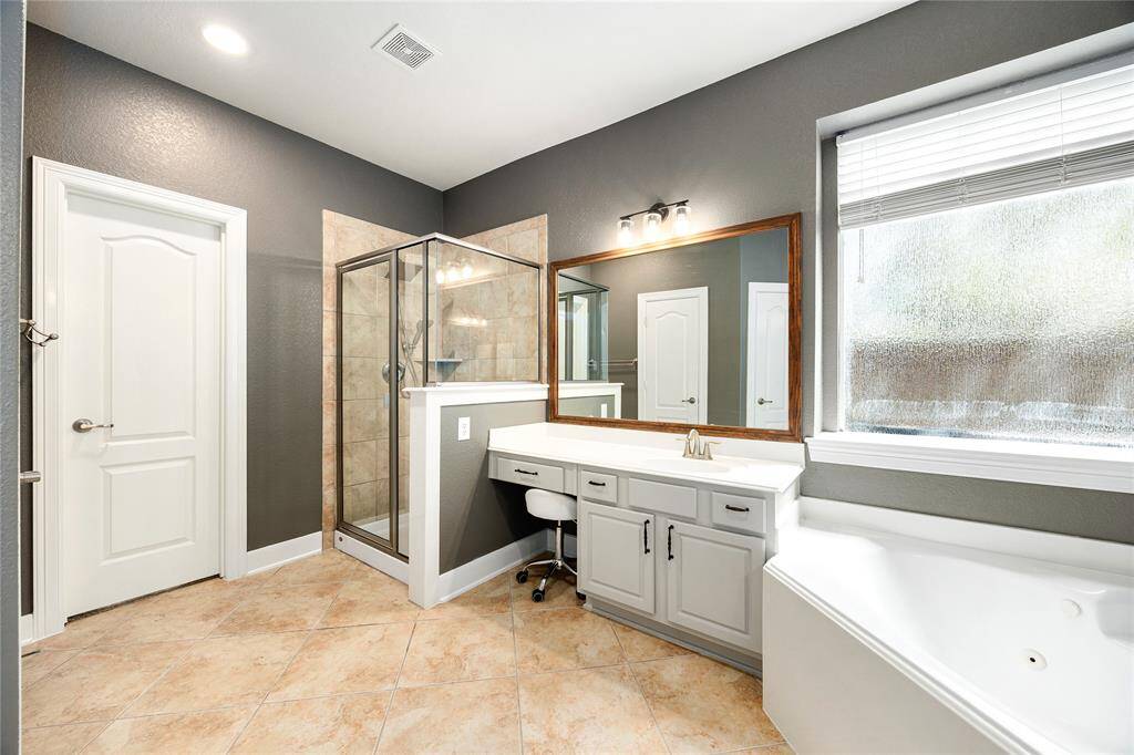 This bathroom features a modern design with a spacious glass-enclosed shower, a large vanity with ample storage, and a soaking tub next to a frosted window for privacy. The neutral tile floor complements the sleek gray walls, creating a clean and inviting space.