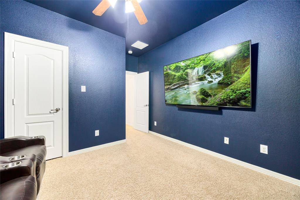 This room features deep blue walls, a plush beige carpet, and a large wall-mounted TV, perfect for a cozy media room. A ceiling fan and white doors add to the modern feel.