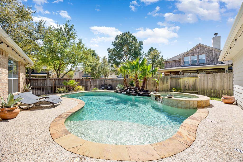 This backyard features a beautiful swimming pool with a stone border, a spa, and a small waterfall. Surrounded by trees and a wooden fence, it offers a private and relaxing outdoor space perfect for enjoying the sun.