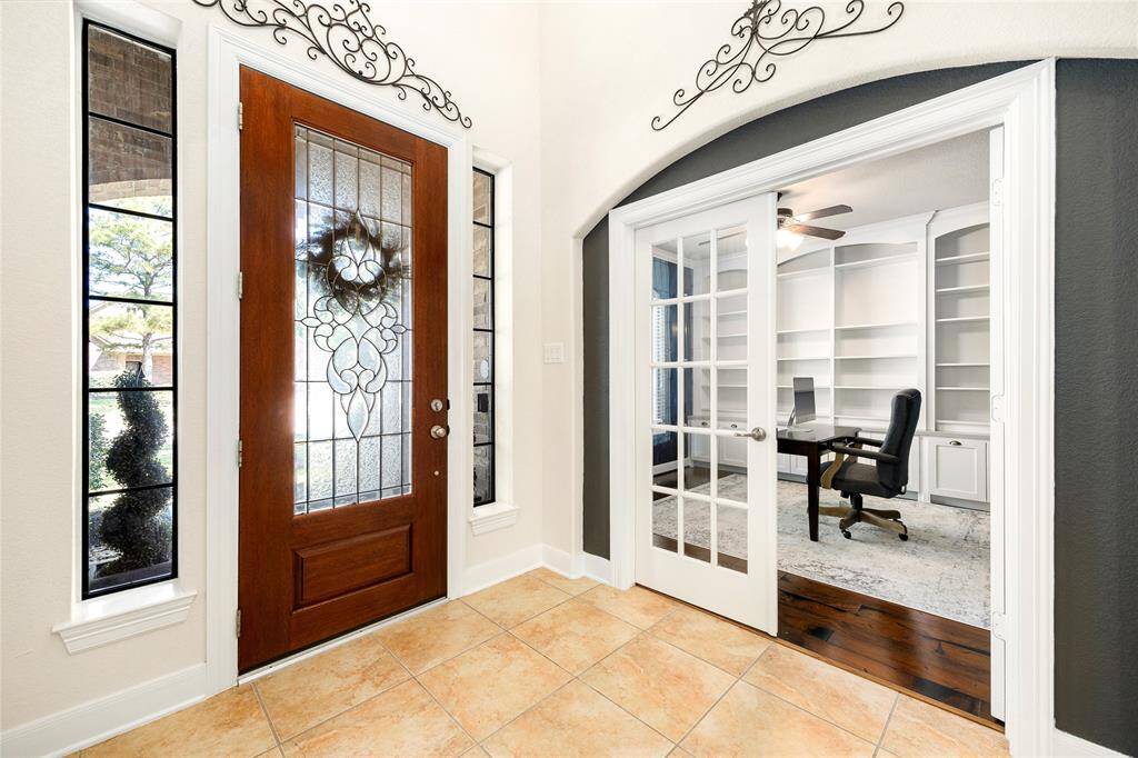 This photo showcases a decorative glass front door and sidelights. It leads to a home office with elegant French doors, built-in shelving, and a ceiling fan, offering a welcoming and functional space.