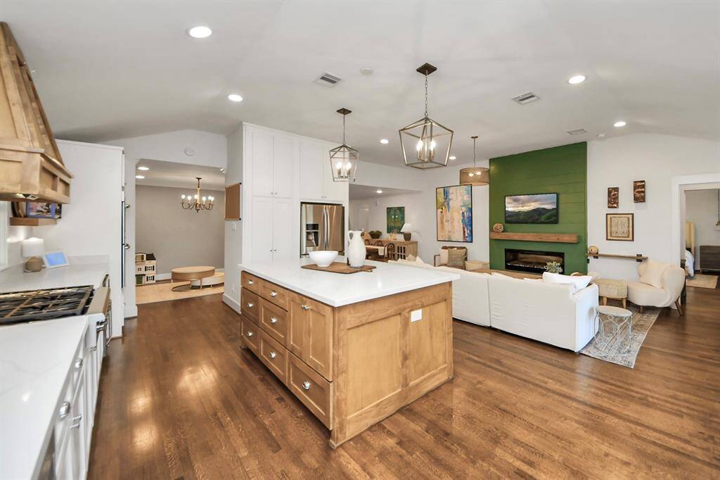 Experience the perfect blend of style and comfort in this open-concept living area, featuring a modern kitchen with a chic island, stainless steel appliances, and elegant pendant lighting. The living room has raised ceilings, a green accent wall, a stylish electric fireplace, and ample seating for a warm, inviting atmosphere.