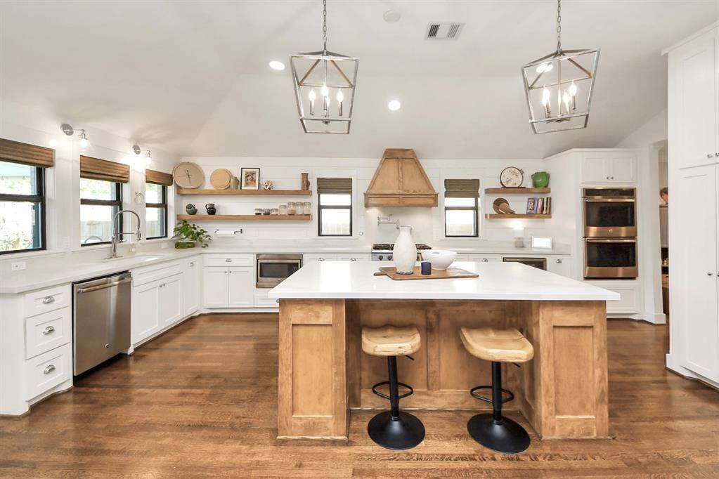 This spacious kitchen features a modern comfortable design with white cabinetry, a large central island with wooden accents, and open shelving. It includes stainless steel appliances, pendant lighting, and ample natural light from multiple windows, creating a bright and inviting space.