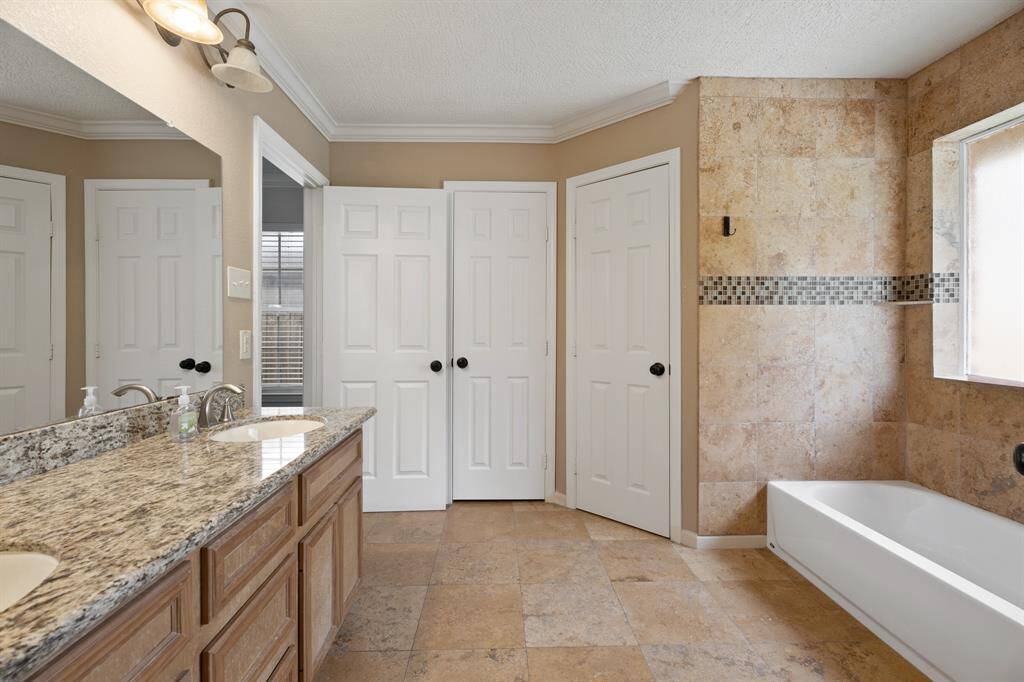 This bathroom features a dual-sink vanity with granite countertops, a large mirror, and warm-toned tile flooring. It includes a bathtub with tiled walls and modern fixtures, providing a spacious and elegant feel.