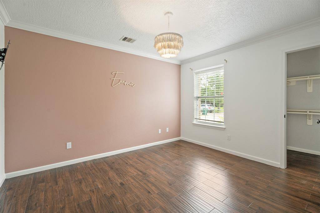 A cozy bedroom with a warm accent wall and wood-style flooring. It features a window for natural light, a small chandelier, and an open closet space, creating a welcoming atmosphere.