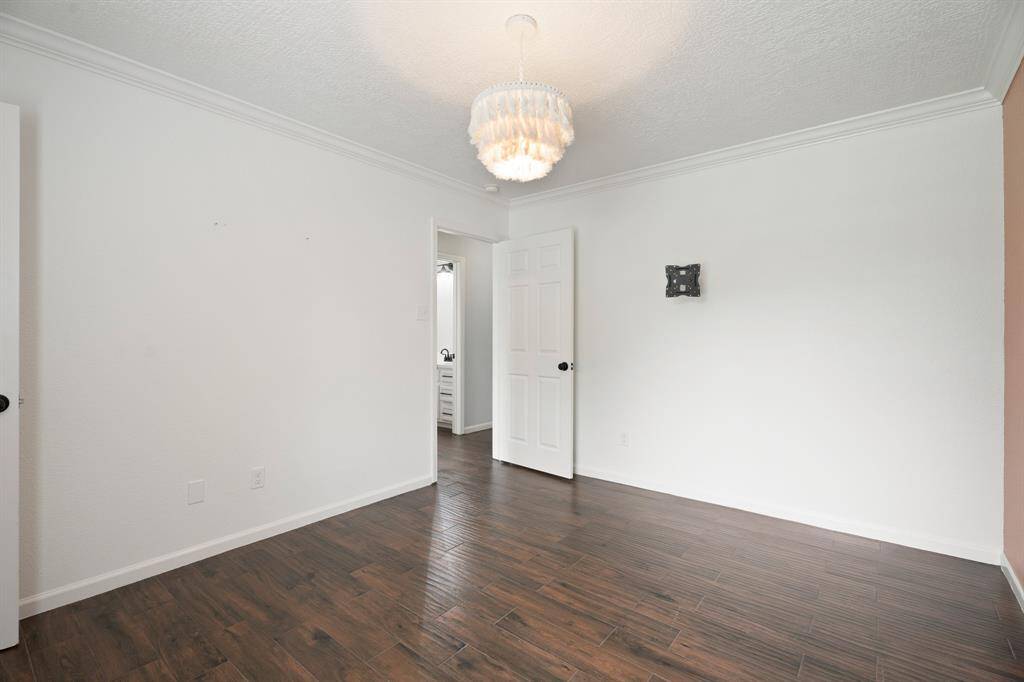 This room features hardwood flooring, a white ceiling with decorative molding, and neutral walls. It includes a hanging light fixture and a door leading to another area of the home. Perfect for a bedroom or office space.