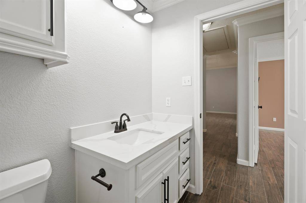 This photo shows a modern bathroom with a sleek white vanity featuring black fixtures and ample storage. It has a well-lit, clean design with light-colored walls and access to other rooms through a doorway with wood-look flooring.