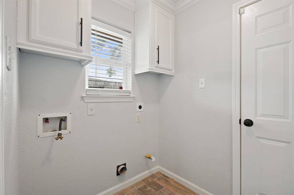 This photo shows a bright, well-organized laundry room with white cabinetry, a window for natural light, and connections for a washer and dryer. The space has a modern feel with light-colored walls and wood-style flooring.