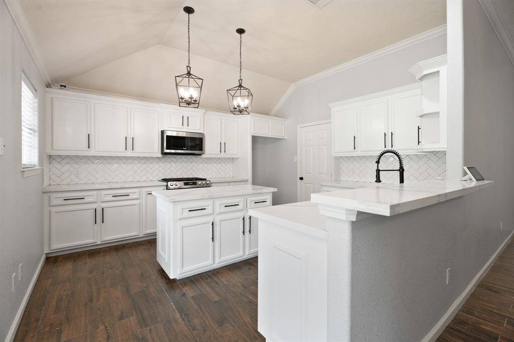 This kitchen features a modern design with white cabinetry, a stylish herringbone tile backsplash, and sleek stainless steel appliances. The space is well-lit with elegant pendant lights and has a central island with ample storage. The dark wood flooring contrasts beautifully with the white decor, creating a warm and inviting atmosphere.