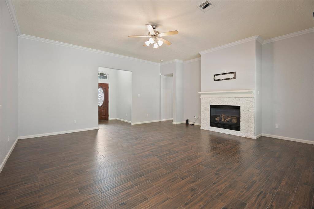 This spacious living room features elegant wood flooring, a cozy fireplace with a white stone surround, and soft gray walls. A ceiling fan provides comfort, and there's a doorway leading to the front entrance with a decorative glass door.