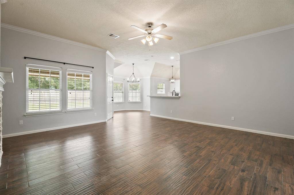 Spacious living area with hardwood floors, neutral wall tones, and crown molding. Features a ceiling fan and large windows for natural light. Open layout flows into a bright dining area and kitchen, ideal for entertaining.