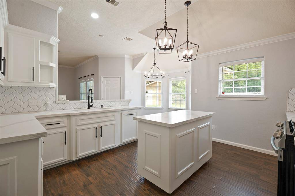 This kitchen features elegant white cabinetry, a central island, and modern pendant lighting. The space is bright with natural light from multiple windows, complemented by a herringbone tile backsplash and dark wood flooring, creating a stylish and functional area.