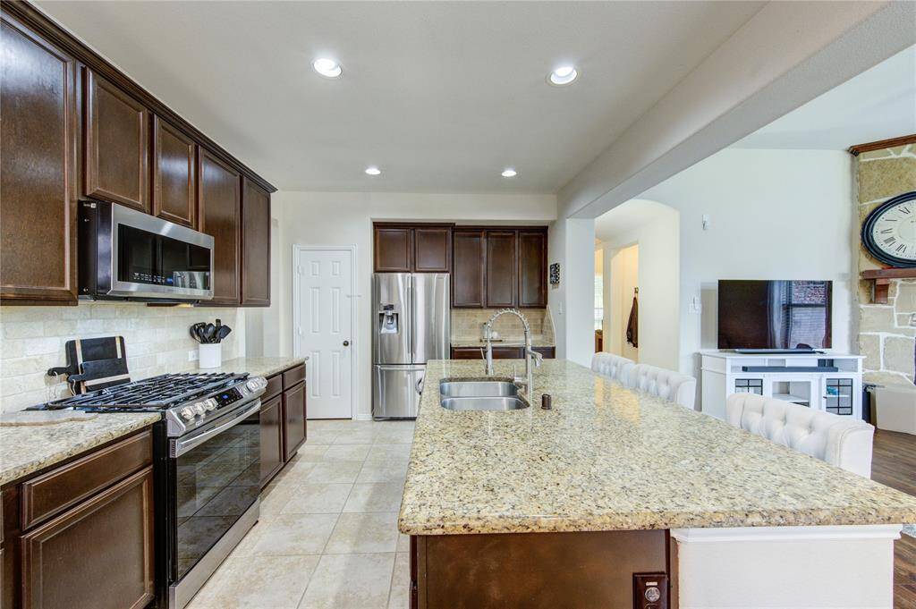 This kitchen features granite countertops, dark wood cabinetry, stainless steel appliances, and an island with seating. It opens into a living area with a fireplace and TV, creating a cozy, modern space.