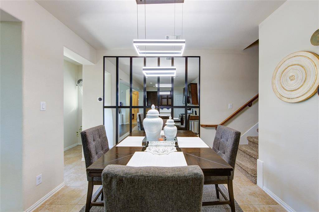 This dining area features a modern design with a sleek table and chairs, accented by contemporary lighting. The space is open and inviting, with a view into the kitchen through stylish glass panels. Neutral tones and decorative wall art create a warm atmosphere, complemented by nearby stairs leading to the upper floor.