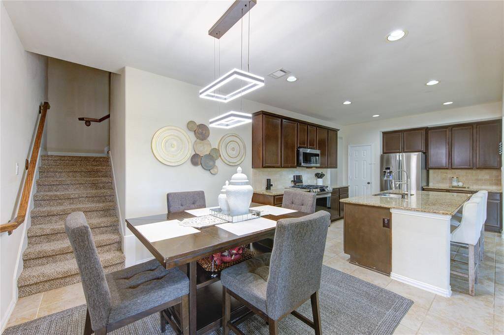 This photo showcases a modern, open-plan kitchen and dining area. The kitchen features dark wood cabinets, stainless steel appliances, and a central island with bar seating. The dining area includes a contemporary table with cushioned chairs, and the space is complemented by stylish lighting fixtures. A carpeted staircase leads to the upper floor, adding a cozy touch.