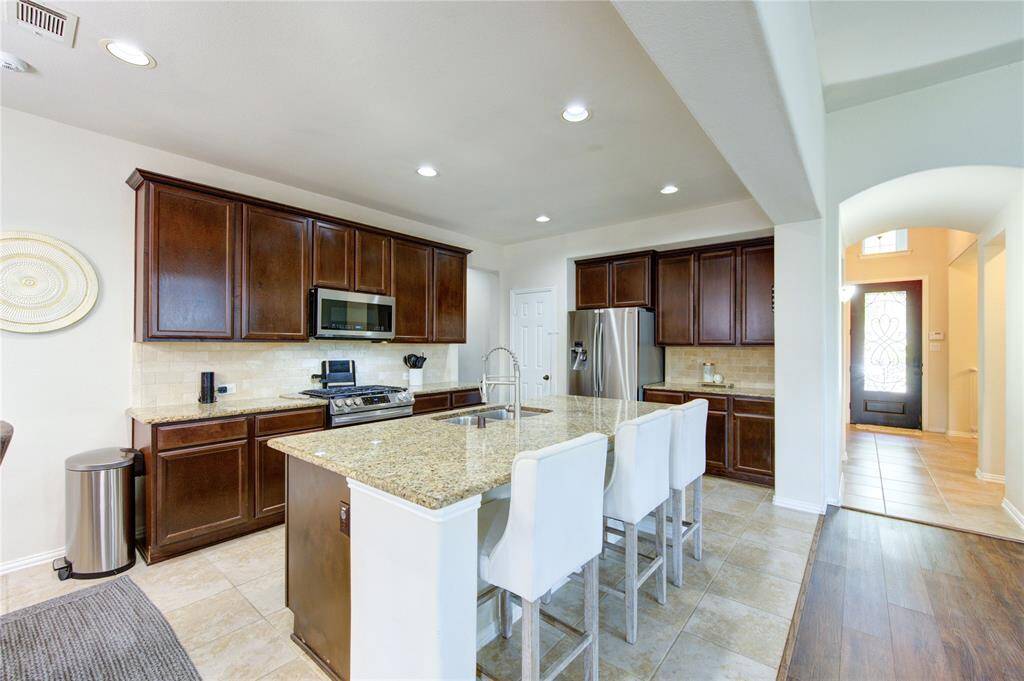 This modern kitchen features granite countertops, dark wood cabinets, stainless steel appliances, and a spacious island with seating. The layout is open, with tile and wood flooring, and it leads into a bright entryway.