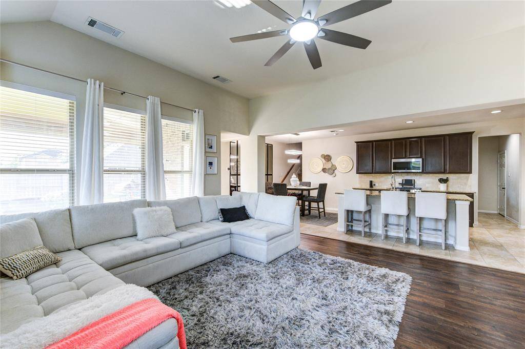 This inviting open-concept living area features a spacious L-shaped sofa, modern ceiling fan, and ample natural light from large windows. The kitchen boasts a breakfast bar with seating, granite countertops, and dark wood cabinetry, seamlessly connecting to a cozy dining area.