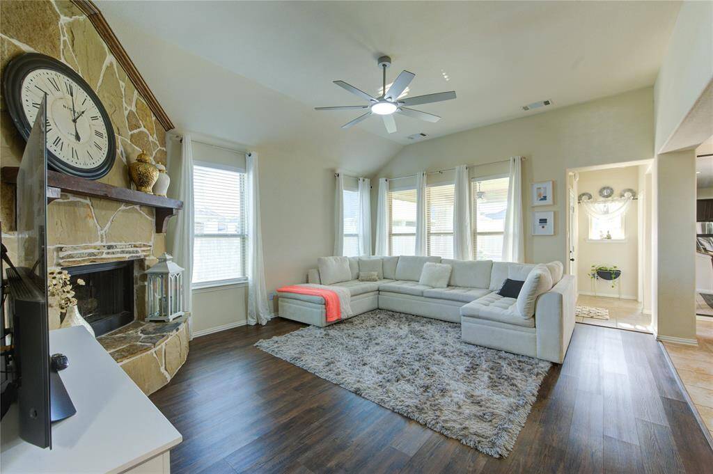 This inviting living room features a high ceiling and abundant natural light, with a cozy stone fireplace and large clock as focal points. A spacious sectional sofa and a plush area rug add comfort, while wood flooring enhances warmth and style.