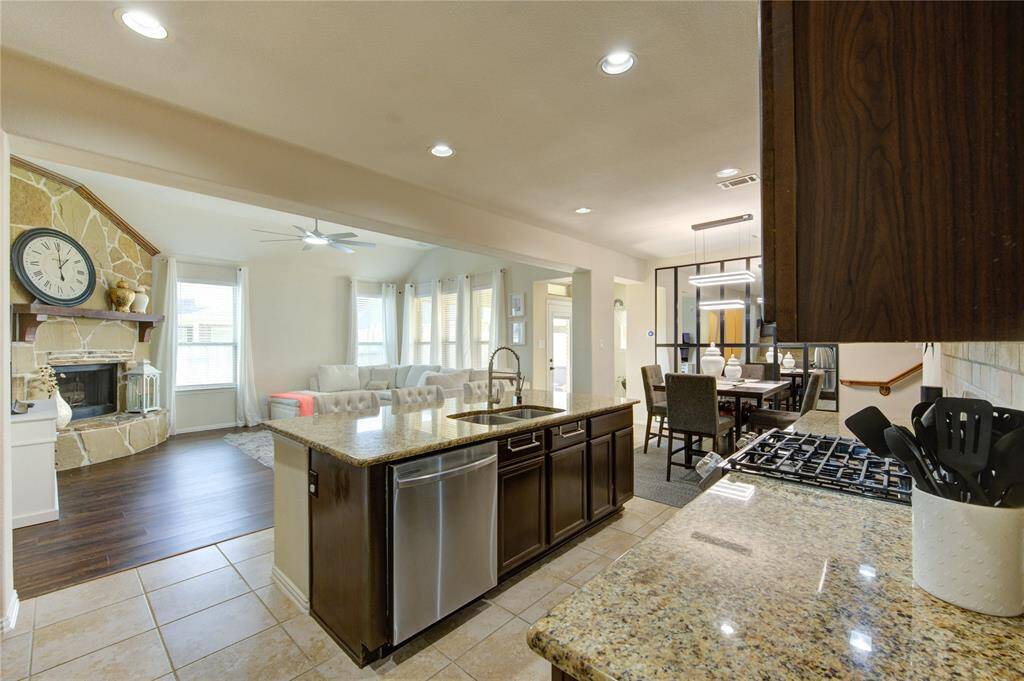 This photo showcases a modern open-concept kitchen and living area. The kitchen features granite countertops, a central island with seating, and stainless steel appliances. The living space has a cozy stone fireplace and large windows for natural light, seamlessly connecting to a dining area with stylish lighting.