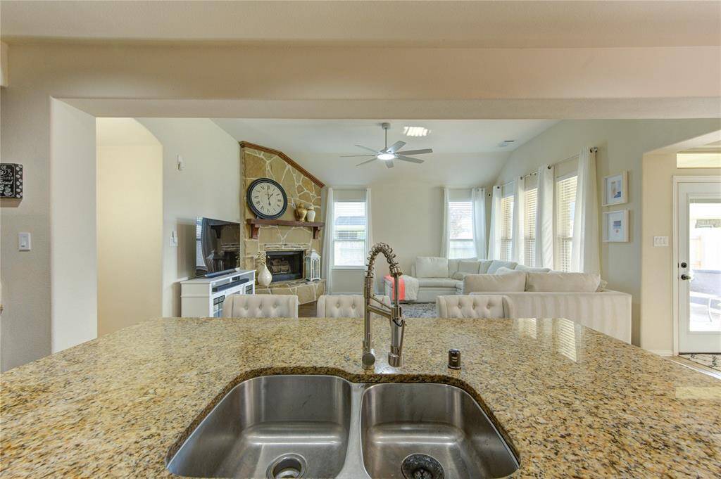 The photo showcases a bright, open living space with a view from a granite kitchen island featuring a double sink. The area includes a cozy stone fireplace, large windows with plenty of natural light, and a comfortable seating arrangement. A ceiling fan adds to the modern, welcoming ambiance.