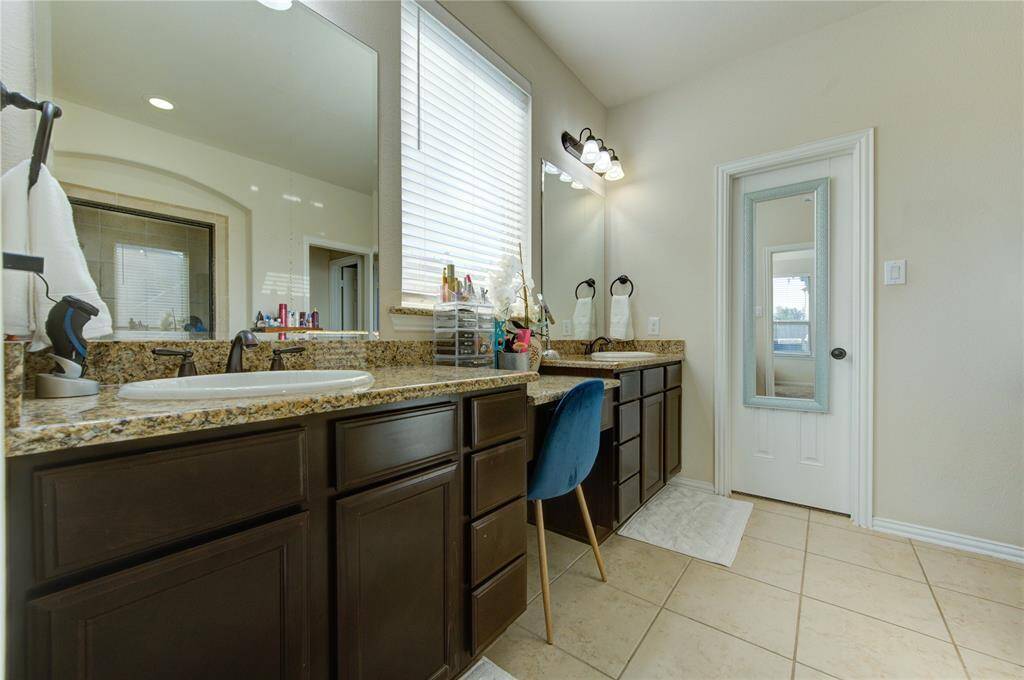 This bathroom offers a spacious layout with dual sinks, granite countertops, and dark wood cabinetry. It features a large mirror, ample natural light, and a separate vanity area with a stylish chair. The neutral tile flooring and modern fixtures add a touch of elegance.