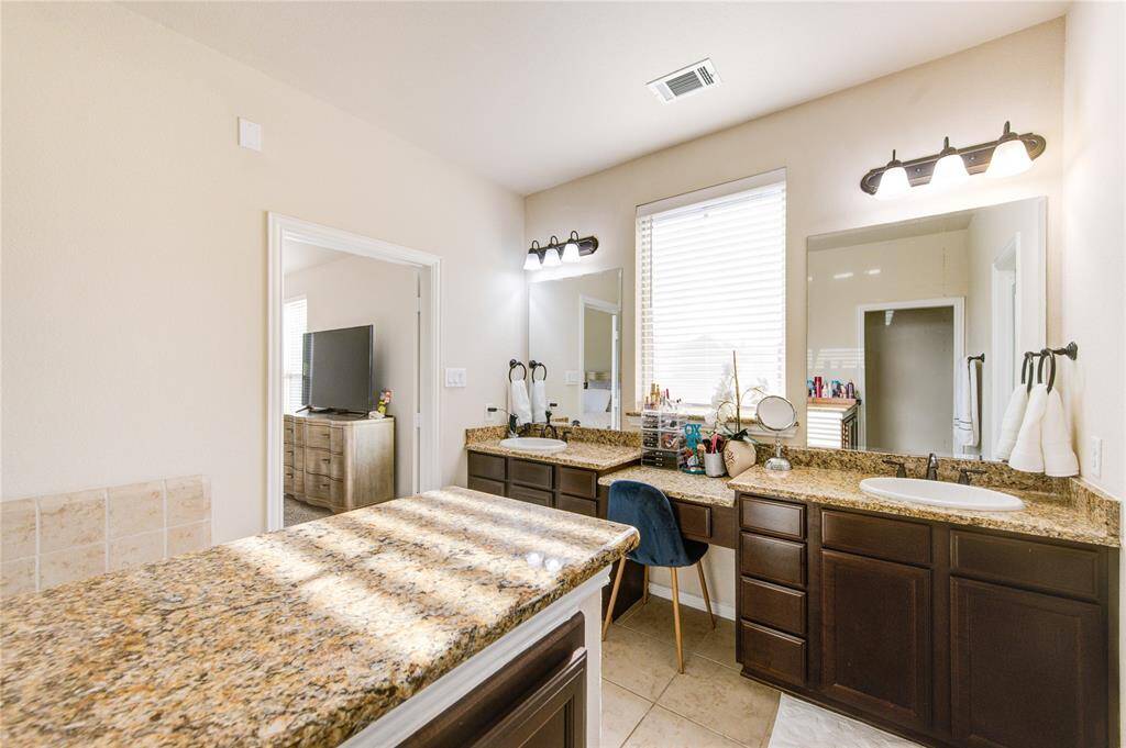 This photo showcases a spacious bathroom featuring dual sinks with granite countertops, dark wood cabinetry, and ample natural light from a large window. A doorway leads to an adjacent room with a TV, enhancing the open and connected layout.