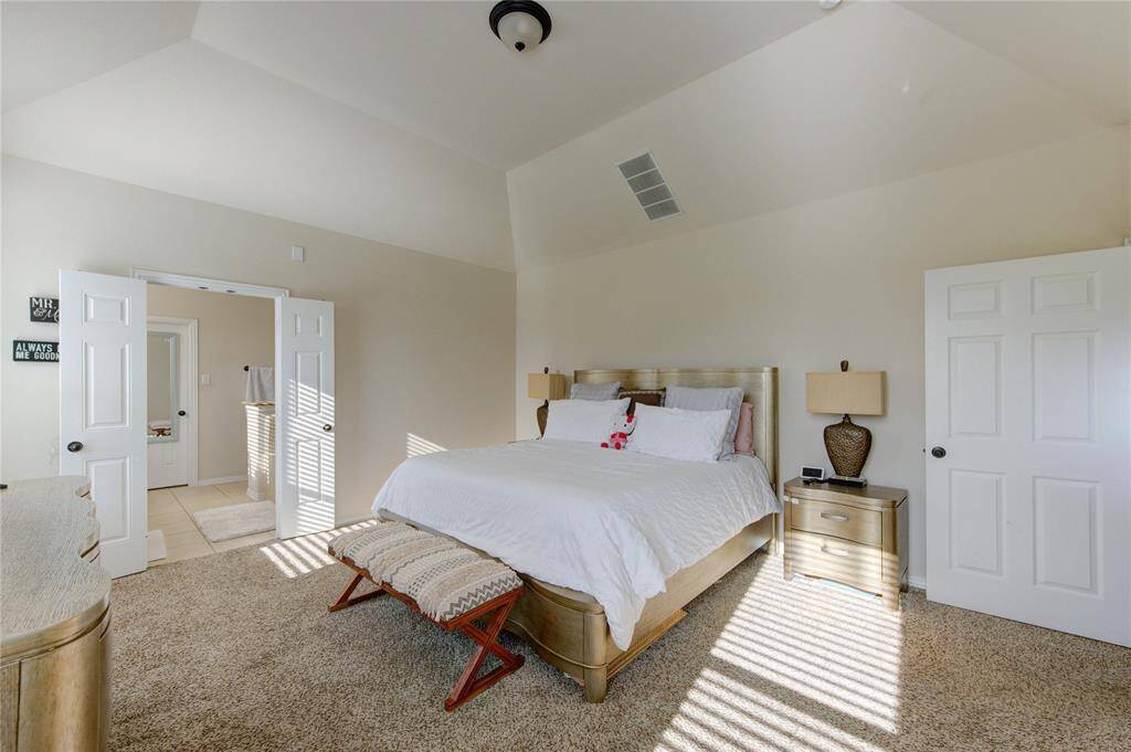 This spacious bedroom features a vaulted ceiling and ample natural light, creating an airy and inviting atmosphere. It includes a large bed with elegant side tables, plush carpeting, and direct access to an en-suite bathroom through double doors, enhancing both comfort and convenience.