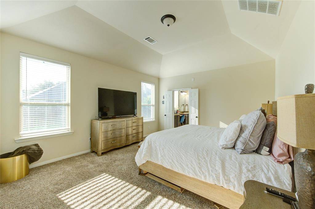 This bright and spacious bedroom features high ceilings, large windows, and carpeted flooring. It includes a comfortable bed, a dresser with a TV, and a view into an en-suite bathroom, creating a cozy and functional retreat.